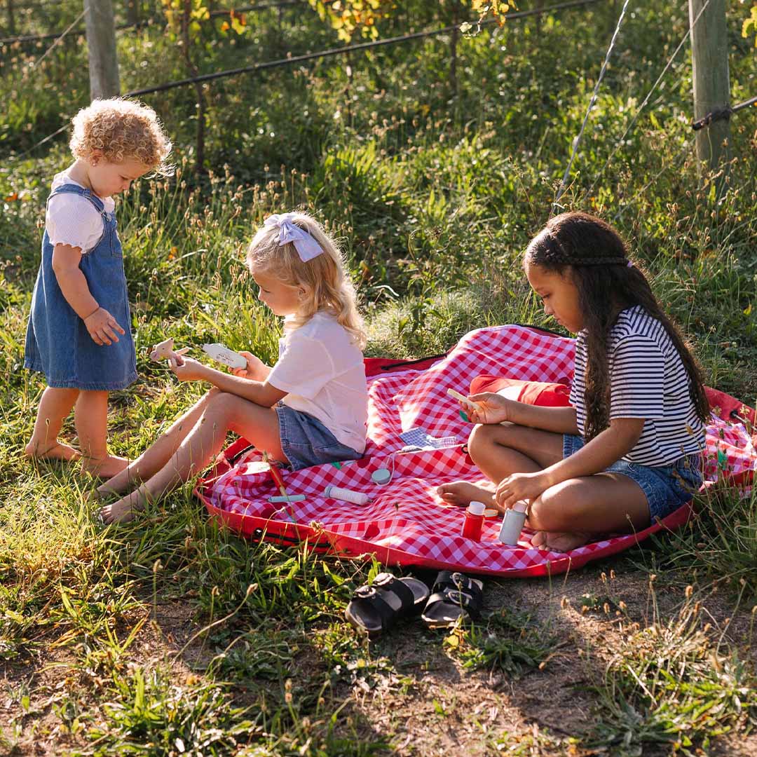 Drie kinderen spelen op een uitgespreid play&go waterdicht speelkleed met rood-wit geruit patroon en kersenmotief in het gras van een wijngaard.