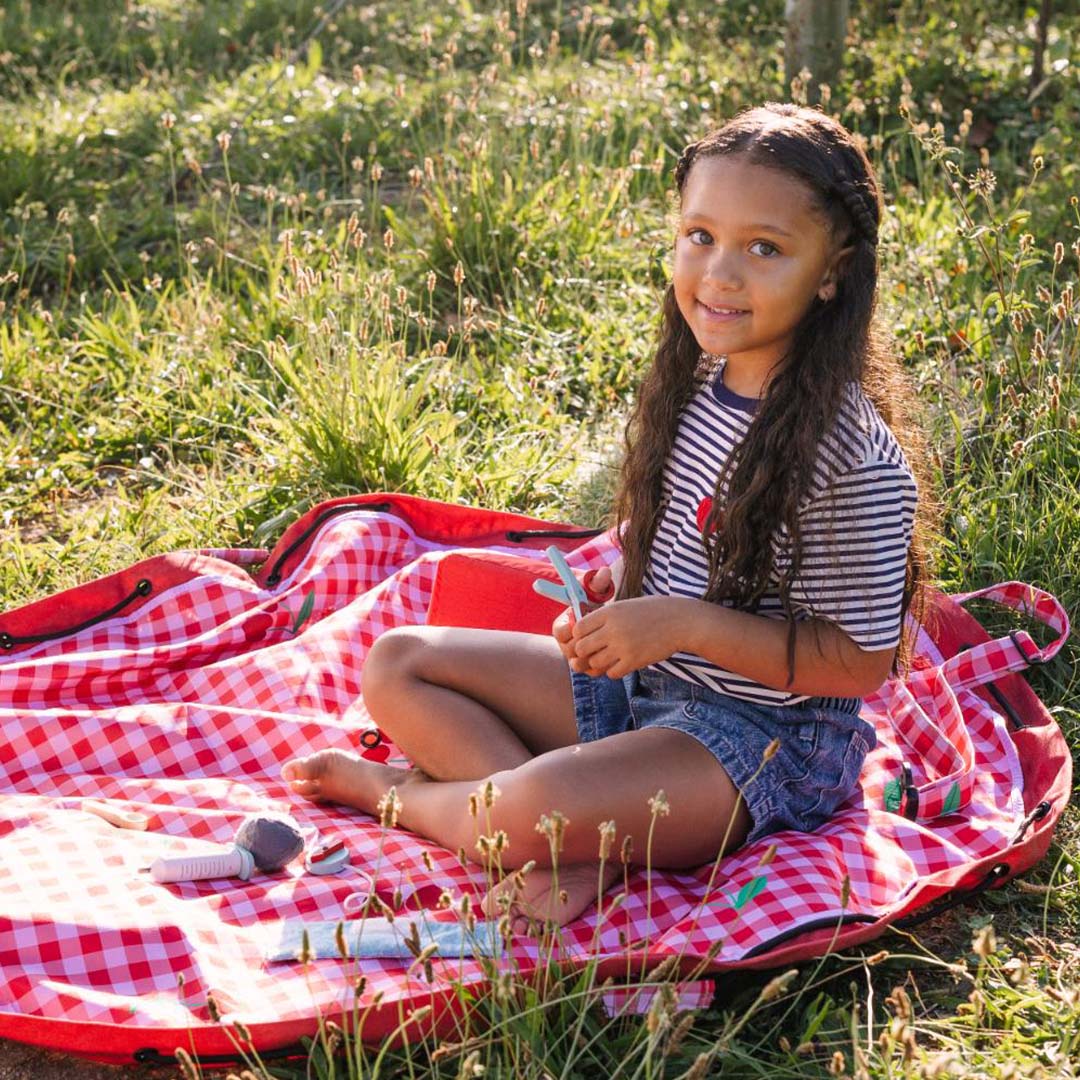 Een lachend meisje zit op een uitgespreid play&go waterdicht speelkleed met rood-wit geruit patroon en kersenmotief in het gras.