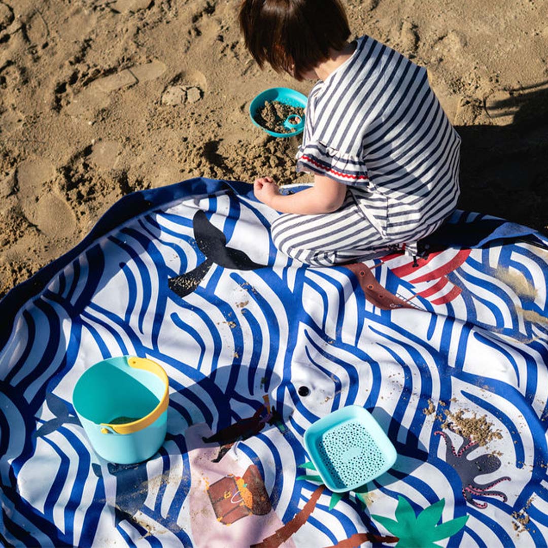 Een kind speelt met zand op het uitgespreide speelkleed met golfpatroon op het strand.