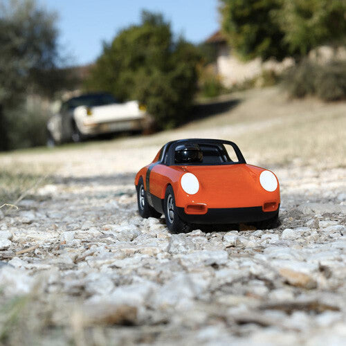 Playforever speelgoedauto Luft Biba in oranje en zwart op een kiezelweg in een landelijke omgeving.