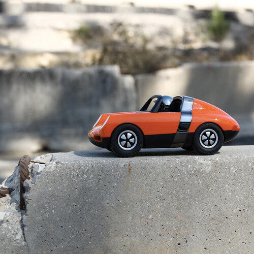 Playforever speelgoedauto Luft Biba in oranje en zwart, van de zijkant gezien op een betonnen muur.
