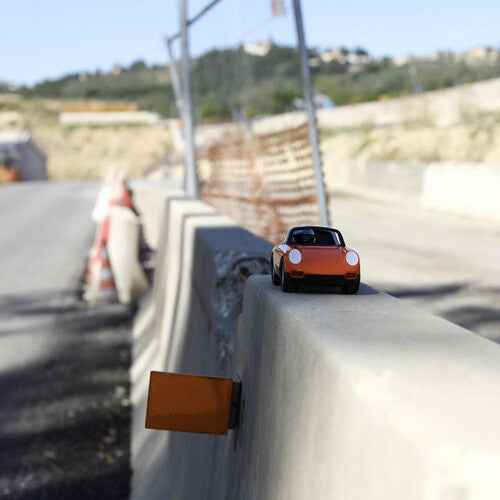 Playforever speelgoedauto Luft Biba in oranje en zwart, van voren gezien op een betonnen vangrail langs een weg.
