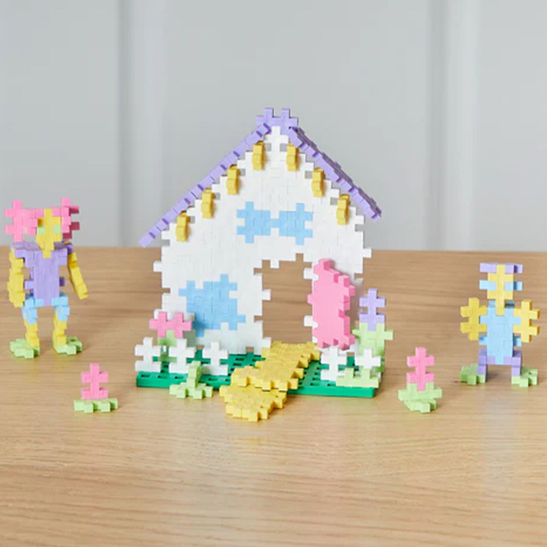 Een huisje en twee figuren gemaakt van pastelkleurige Plus-Plus bouwstenen op een houten tafel.