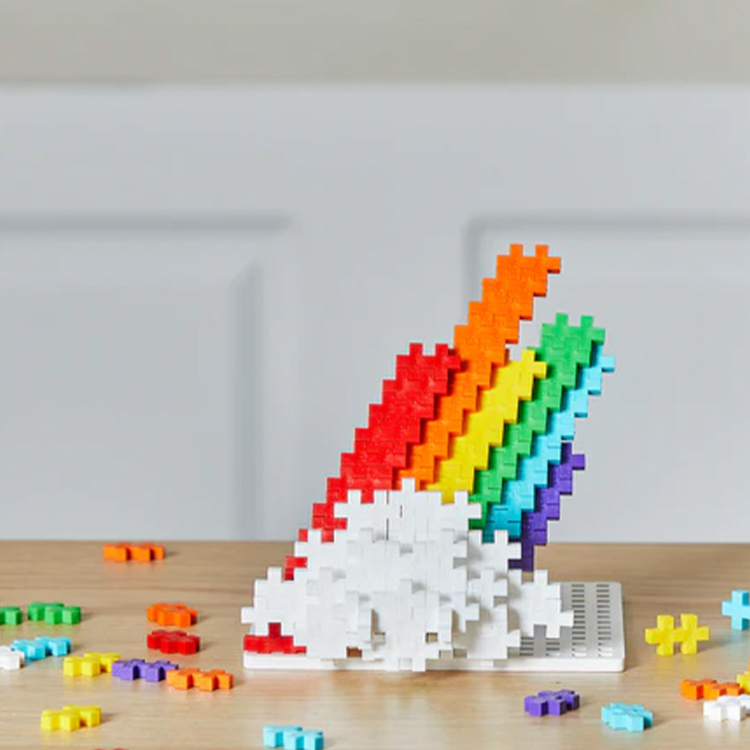 Een regenboogfiguur gemaakt van Plus-Plus bouwblokken op een houten tafel.