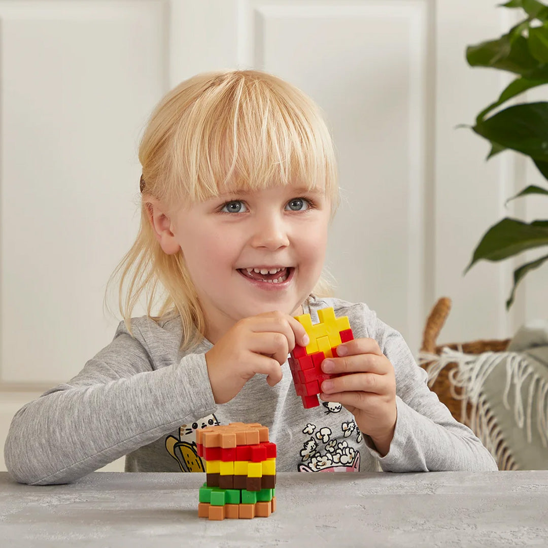 Een lachend meisje houdt een Plus-Plus Big bouwsel van frietjes vast, met een hamburger van bouwstenen op tafel.