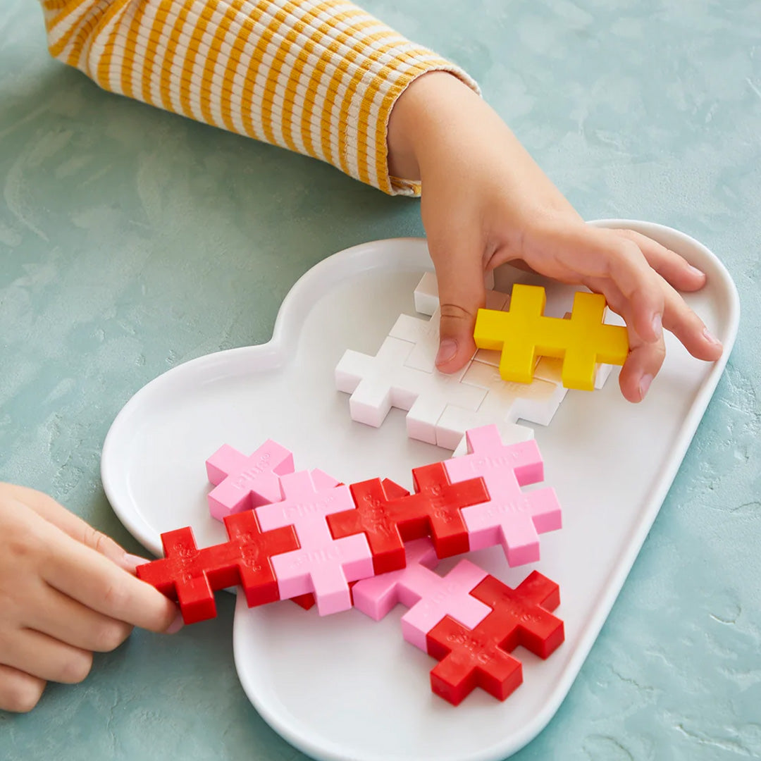 Kinderhanden spelen met Plus-Plus Big bouwstenen in verschillende kleuren op een wit dienblad.