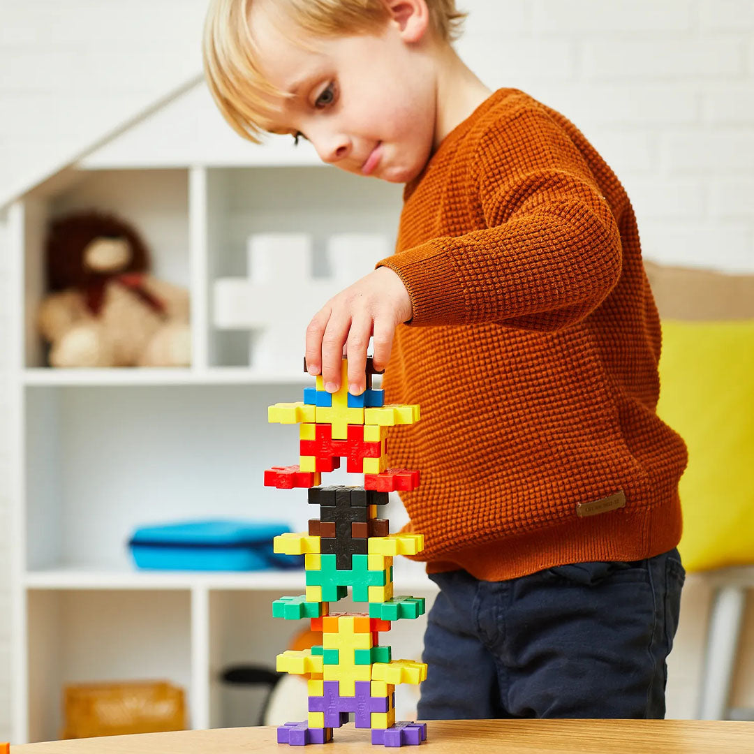 Een jongen bouwt een hoge, kleurrijke toren met Plus-Plus BIG bouwstenen op een houten tafel.