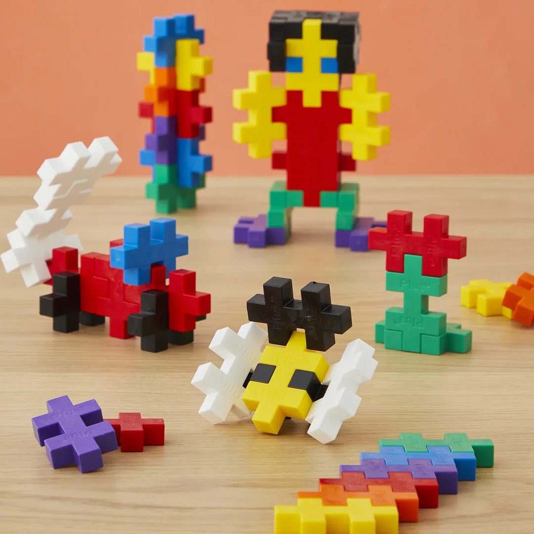 Diverse kleurrijke figuren, waaronder een persoon en een bij, gemaakt van Plus-Plus BIG bouwstenen op een houten ondergrond.