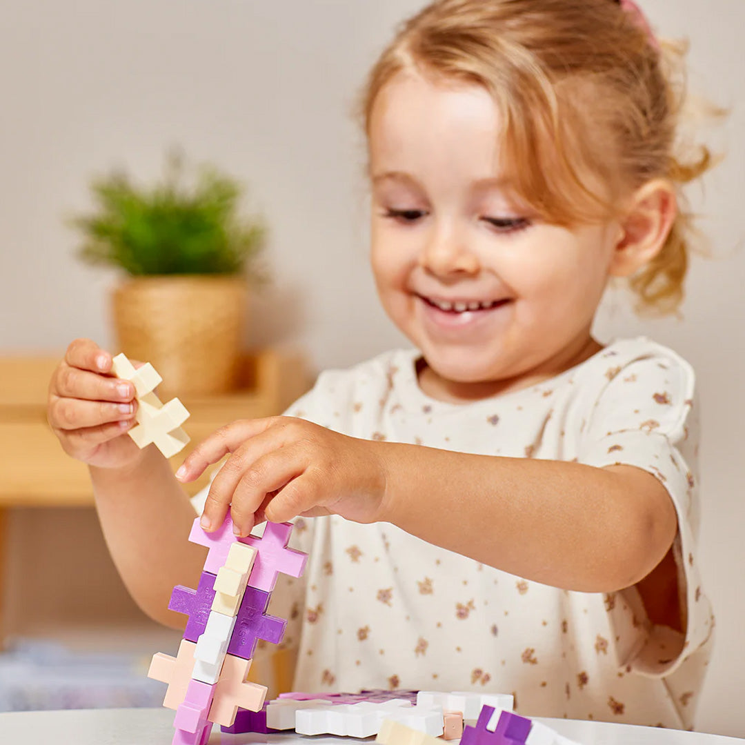 Een lachend jong kind speelt met Plus-Plus BIG bouwblokken in pastelkleuren.