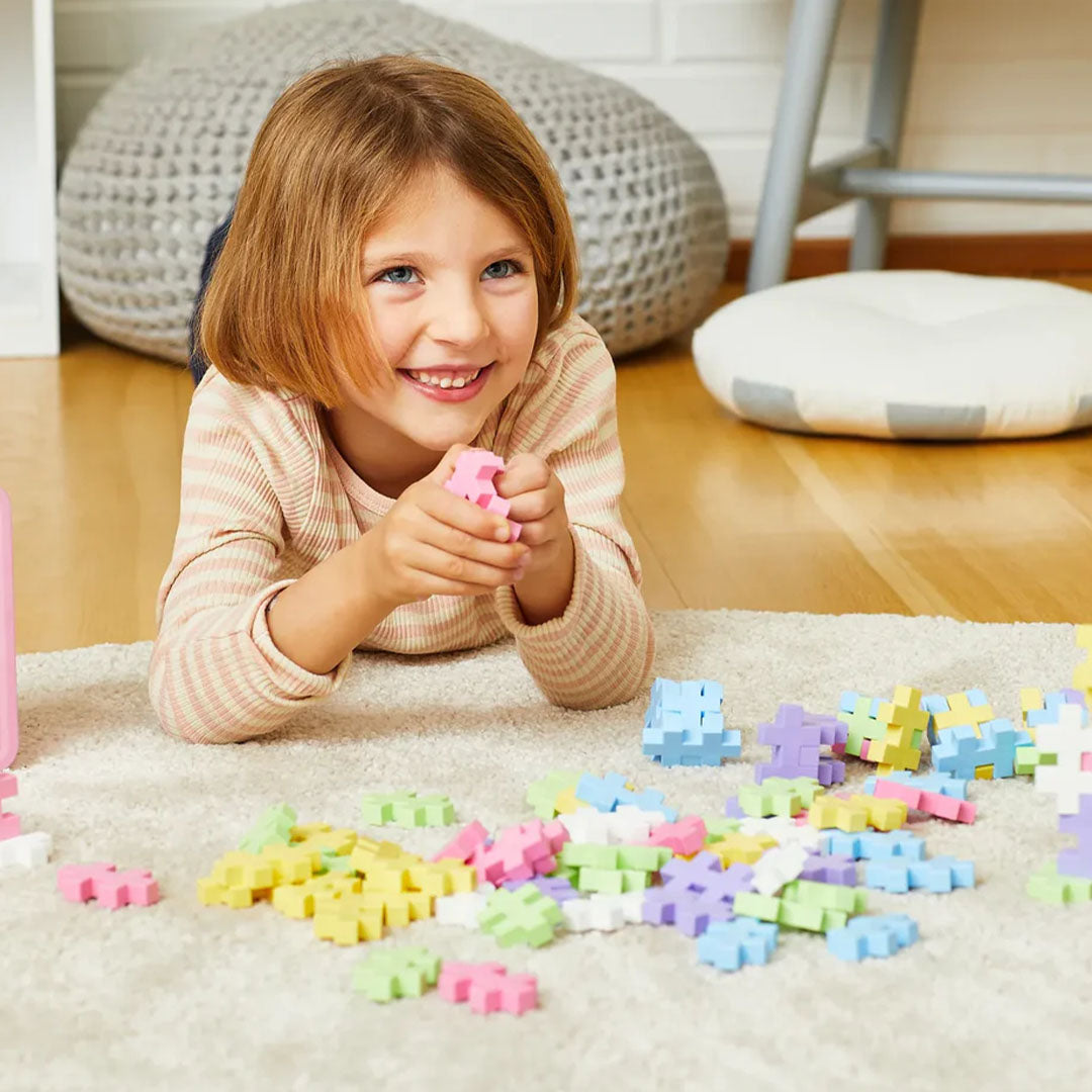 Een lachend meisje speelt op de grond met pastelkleurige Plus-Plus bouwblokken.