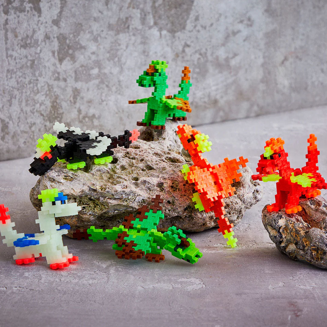 Een collectie van zes kleurrijke drakenfiguren, gemaakt van Plus-Plus bouwstenen, staand op rotsen.