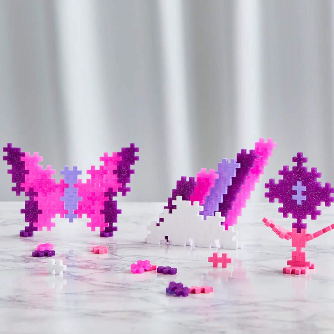 Plus-Plus glitter bouwblokken vormen een vlinder, een regenboog en een figuurtje op een marmeren ondergrond.