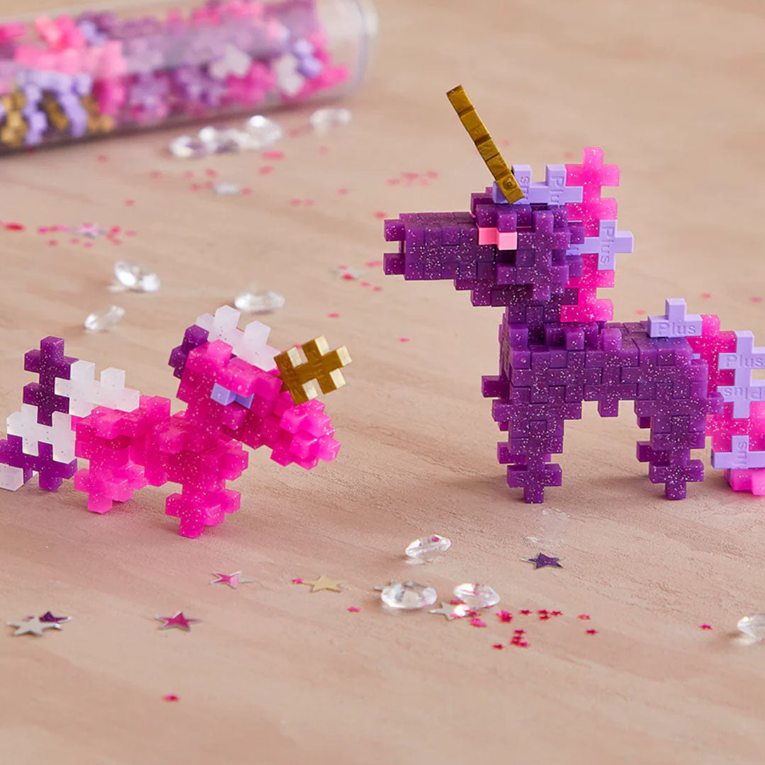 Twee gebouwde Plus-Plus glitter eenhoorns, een paarse en een roze, op een houten ondergrond.