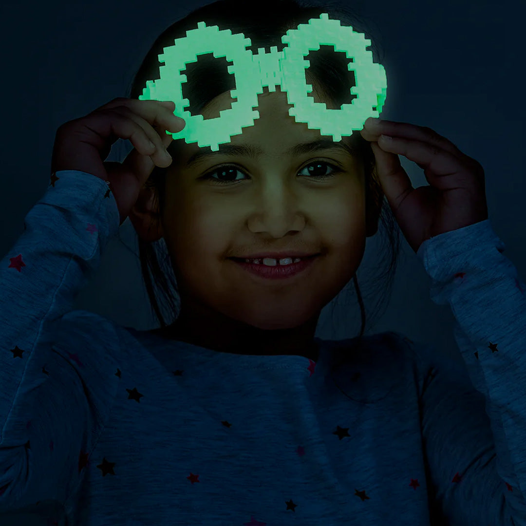 Een lachend meisje draagt een bril gemaakt van glow-in-the-dark Plus-Plus bouwblokken.