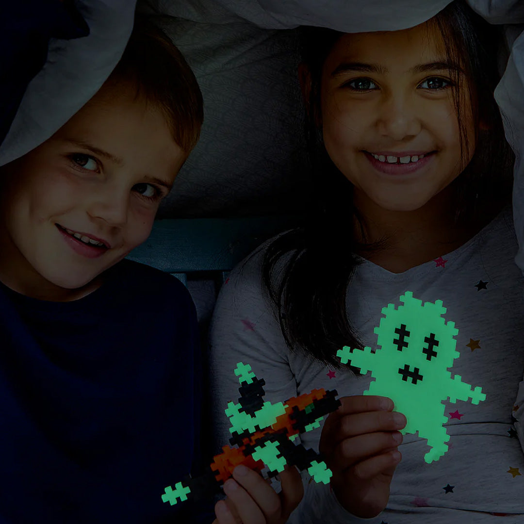Twee kinderen onder een deken spelen met glow-in-the-dark Plus-Plus bouwblokken, een spook en een draak.