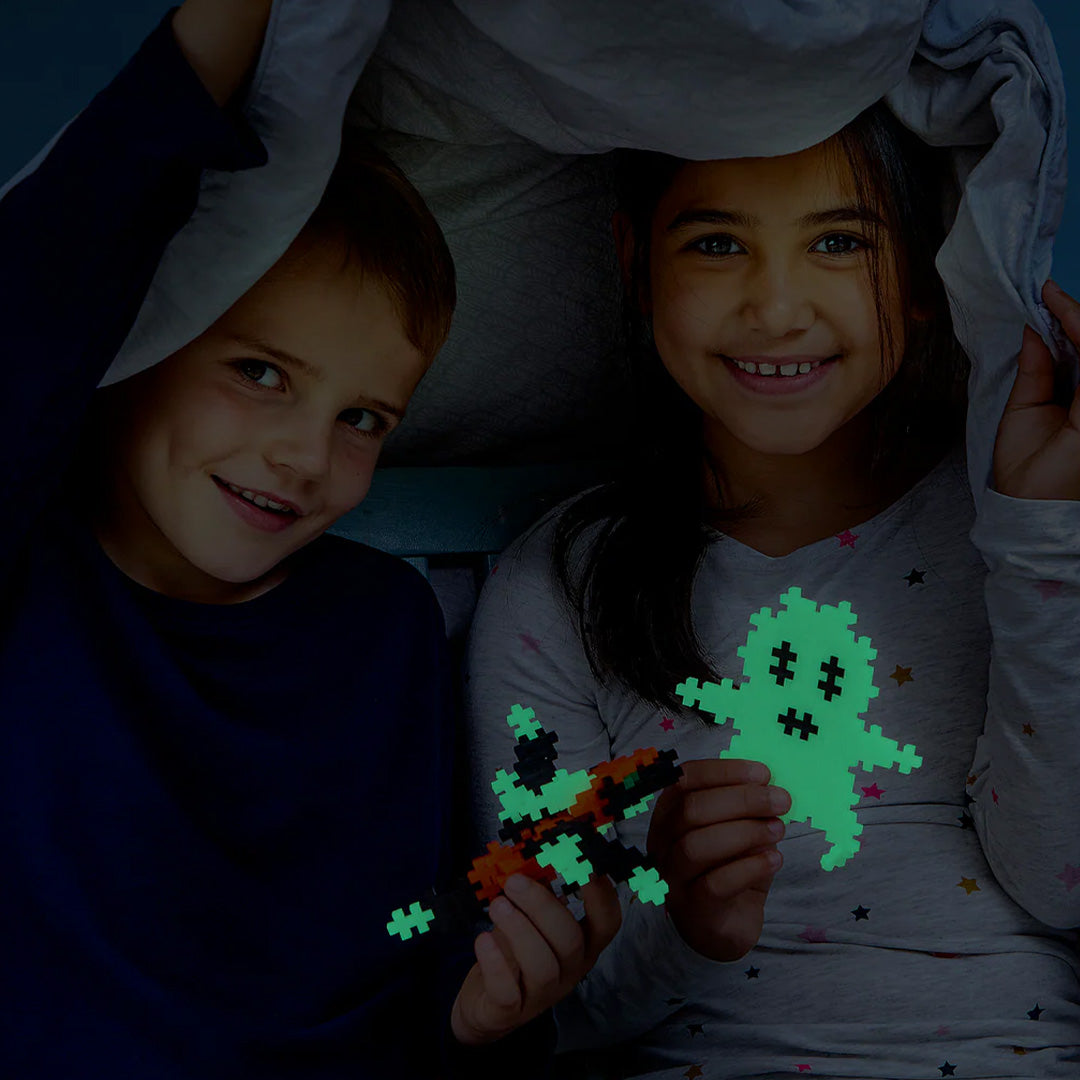 Twee lachende kinderen onder een deken in het donker met lichtgevende Plus-Plus bouwblokken creaties.