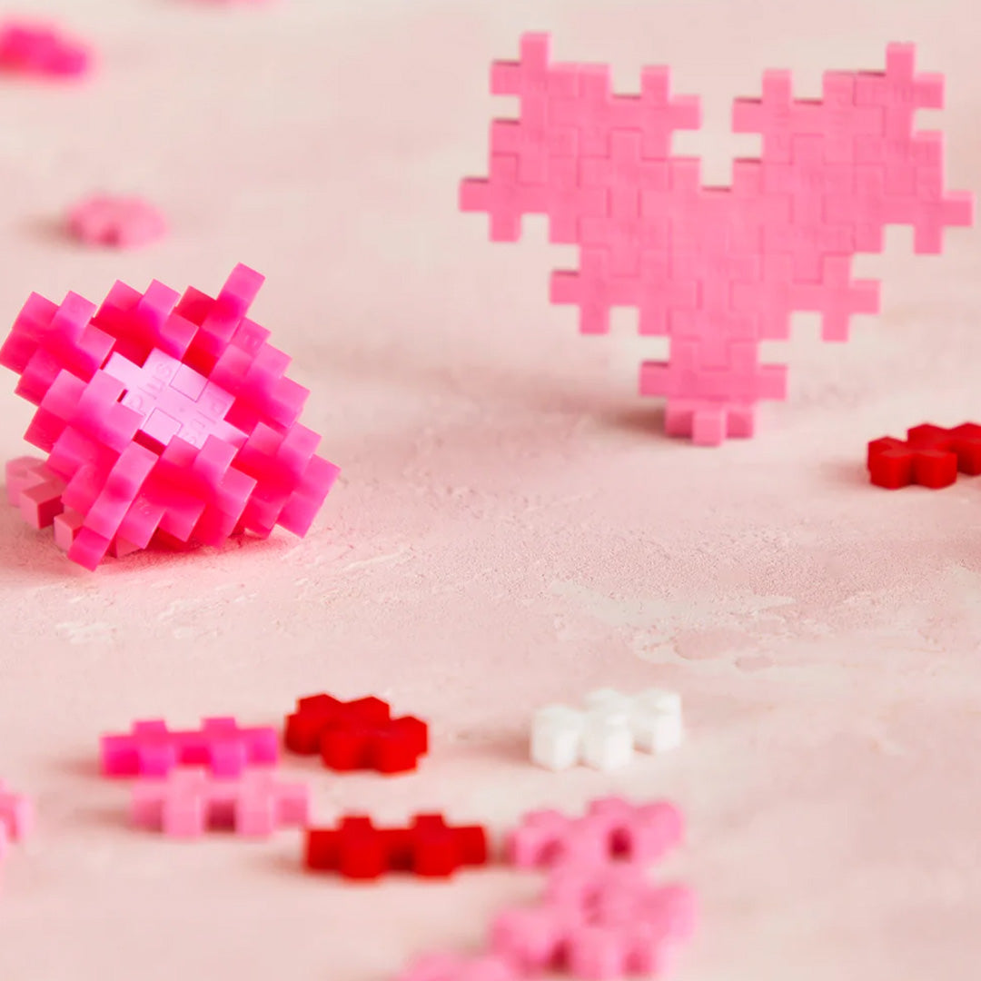 Twee roze harten, gemaakt van Plus-Plus bouwblokken, staan op een roze ondergrond met losse bouwblokken.