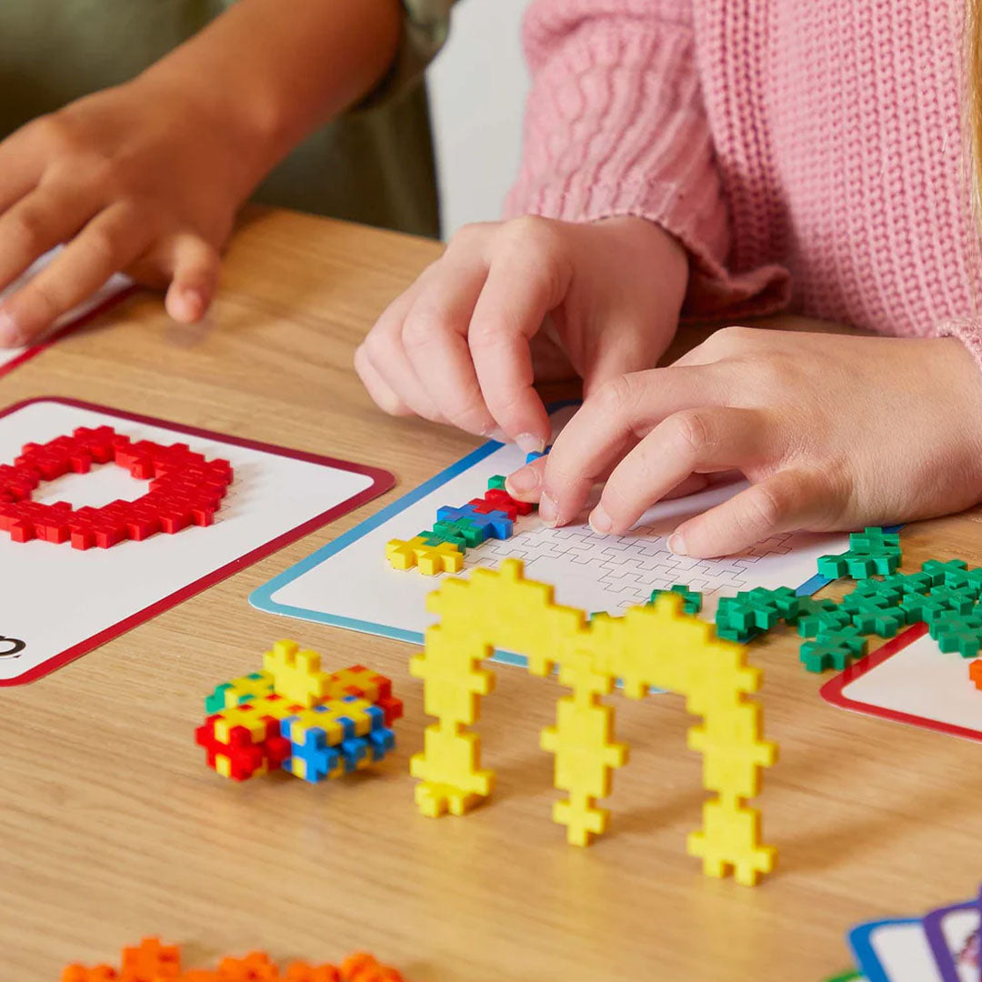 Kinderhanden bouwen met Plus-Plus bouwblokken en instructiekaarten op een houten tafel.