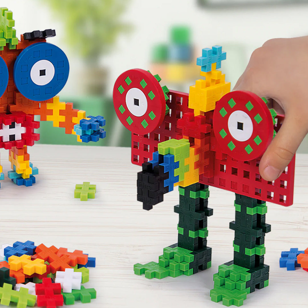 Een hand houdt een groot rood en groen Plus-Plus monsterfiguur vast, met losse bouwblokken op tafel.