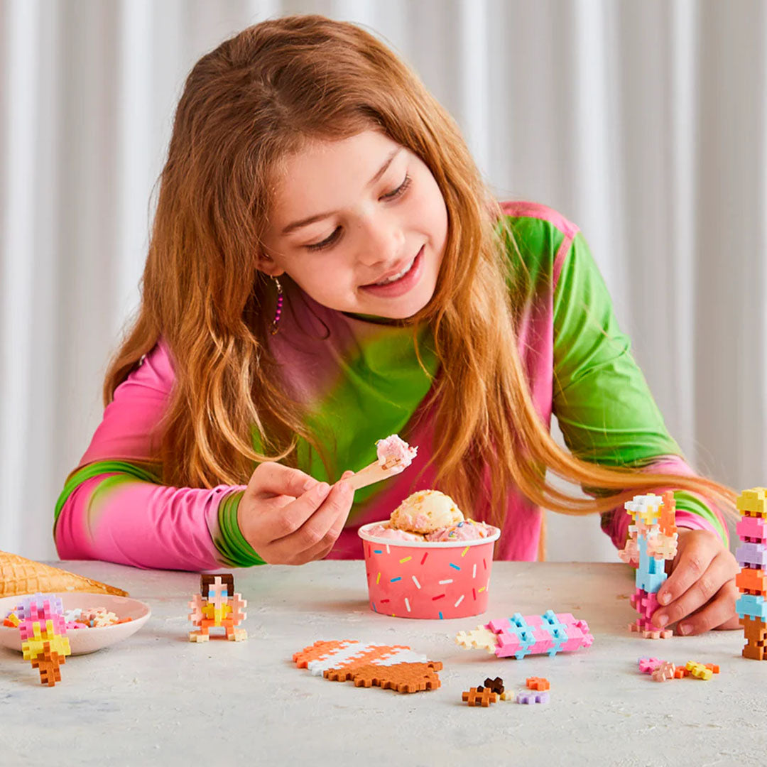Een meisje speelt met Plus-Plus bouwblokken en eet een bolletje ijs uit een roze beker.