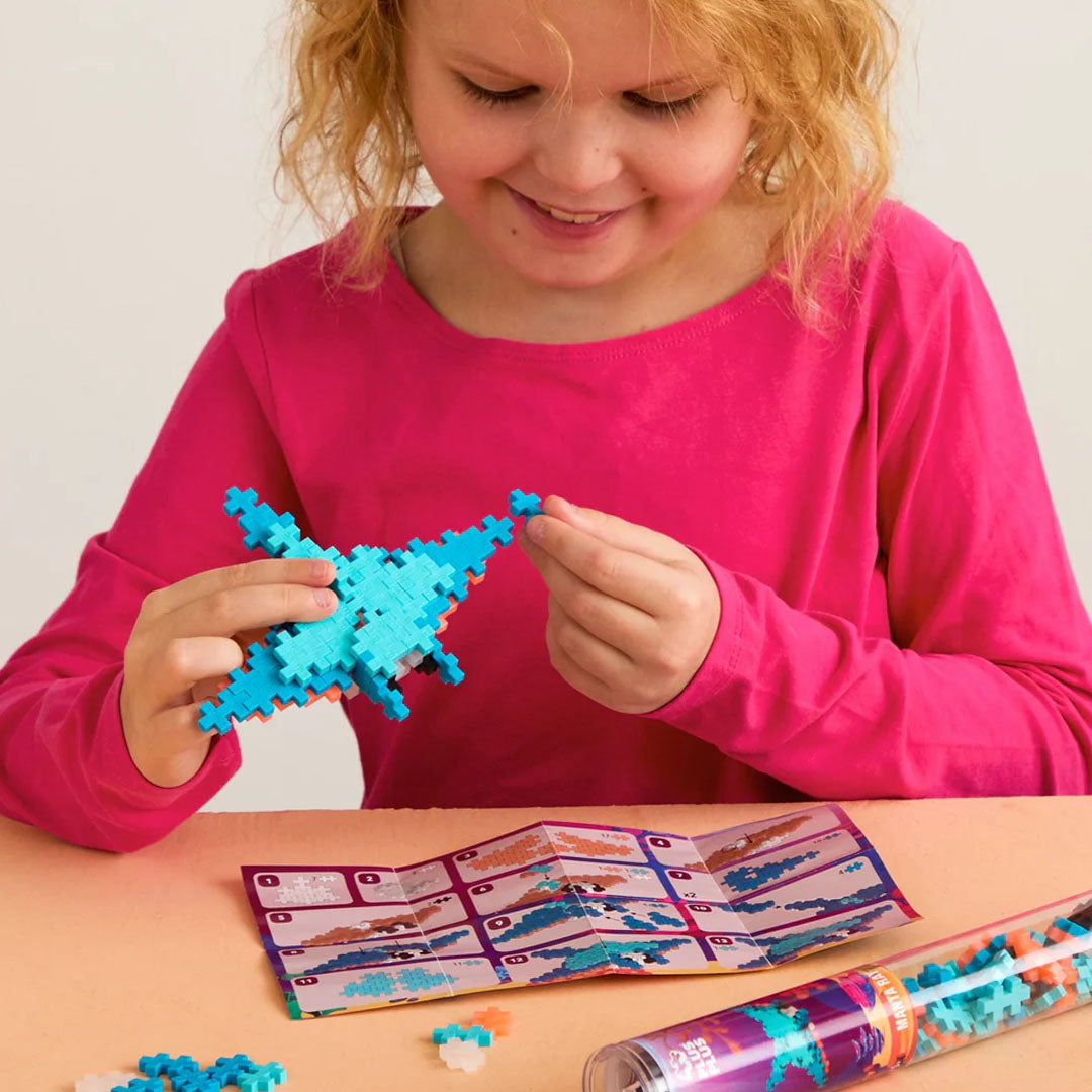 Een lachend kind bouwt een manta ray van Plus-Plus bouwblokken aan tafel met instructies en de verpakking ernaast.