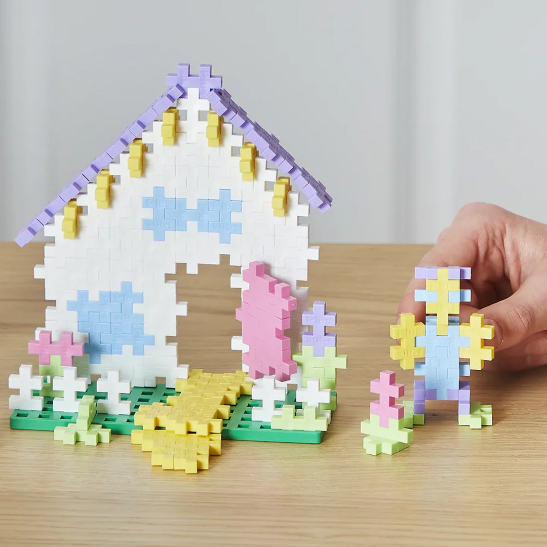 Een huisje en een figuur gemaakt van Plus-Plus pastel bouwblokken staan op een tafel, met een hand die een blokje vasthoudt.