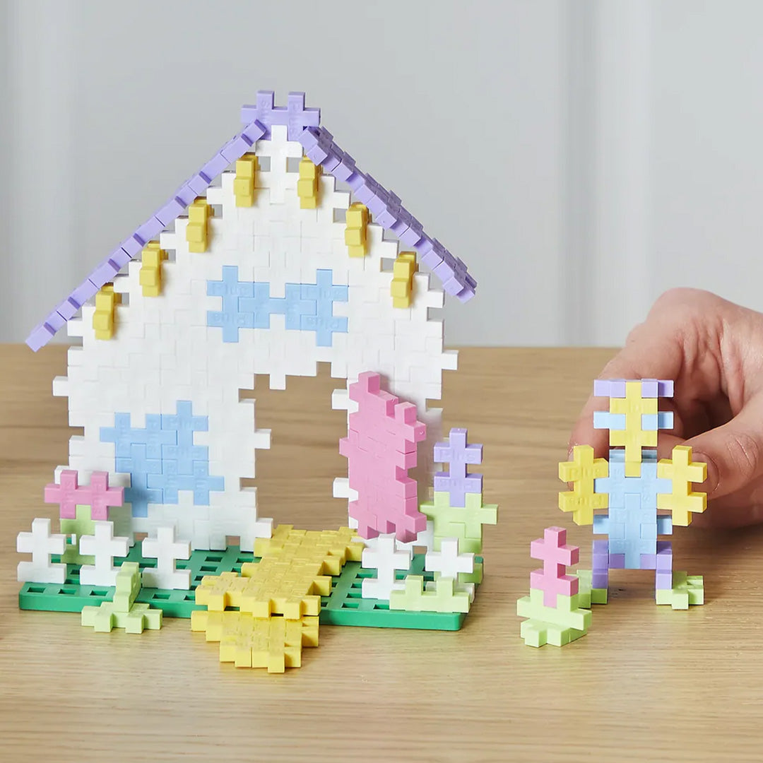 Een huisje en een figuur gebouwd van Plus-Plus bouwblokken in pastelkleuren op een houten tafel.