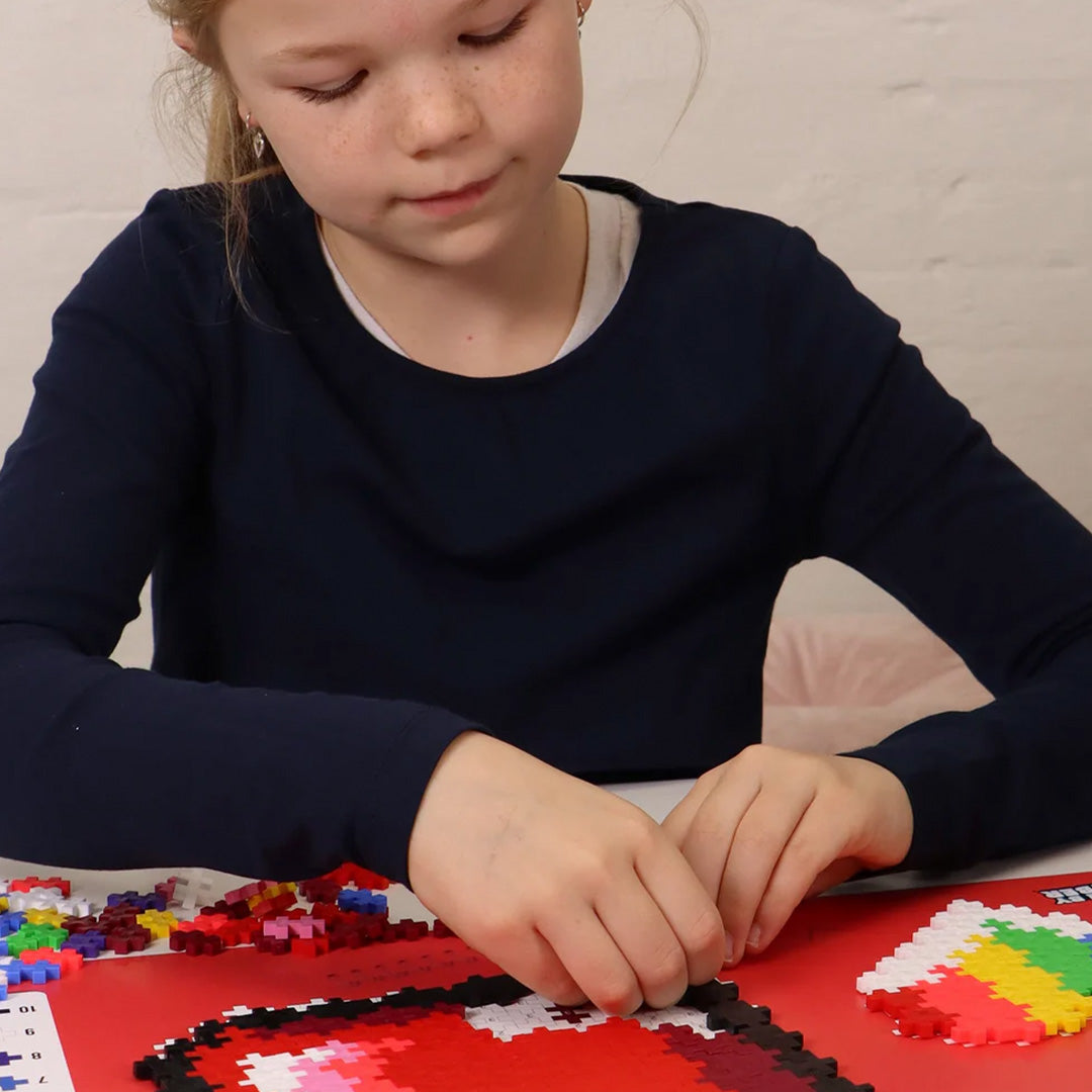 Een kind bouwt een groot rood hart met Plus-Plus bouwblokken op een rode mat.