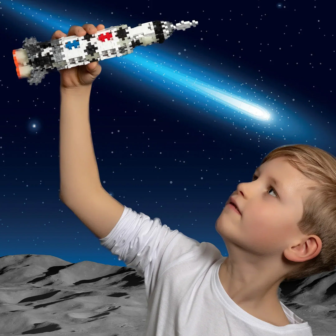 Een kind houdt een gebouwde Saturn V raket van Plus-Plus bouwstenen omhoog tegen een sterrenhemel.