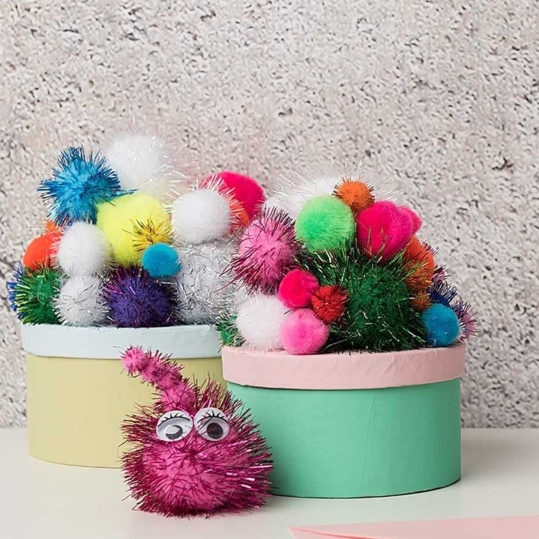 Knutselwerk met veelkleurige glitter pompons die twee ronde dozen versieren en een roze pompon met ogen.