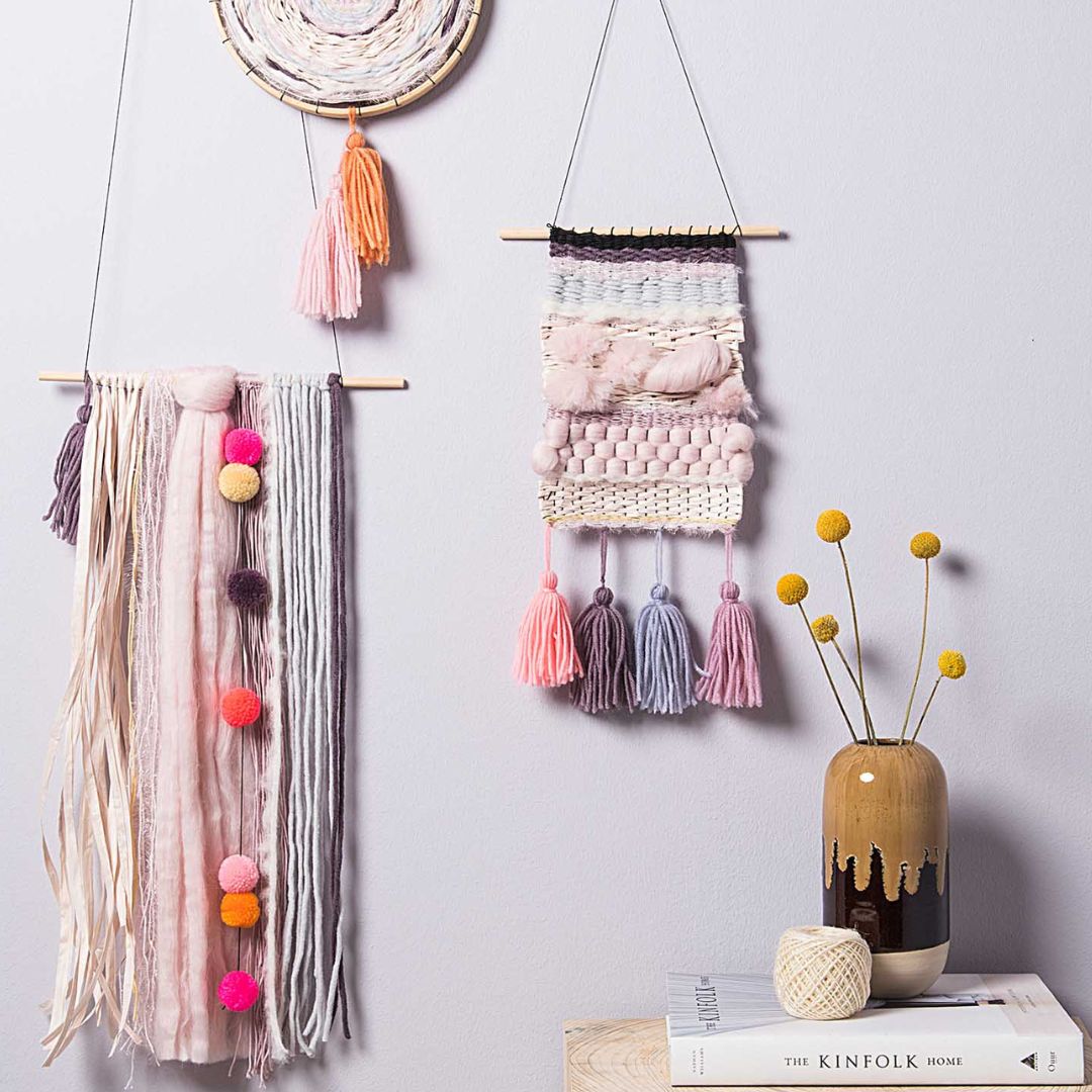 Twee wanddecoraties gemaakt van garen en pastelkleurige pompons hangen aan een grijze muur.