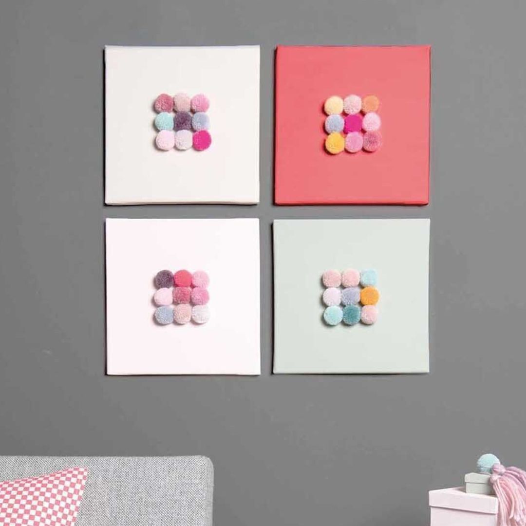 Vier vierkante schilderijtjes aan een grijze muur, elk versierd met een groepje veelkleurige pompons.