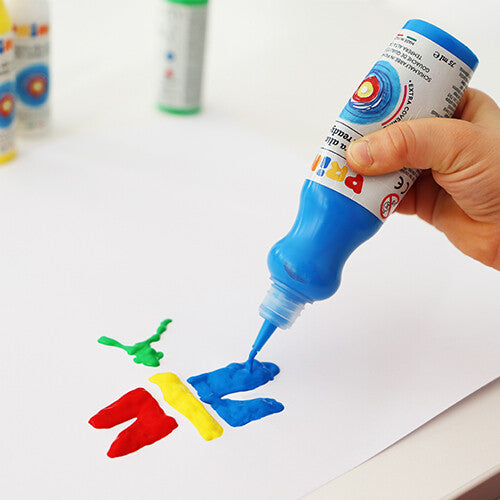 Een hand knijpt Primo blauwe plakkaatverf uit een flesje op wit papier, naast rode en gele verf.