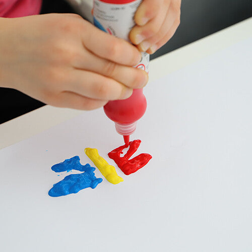 Een kinderhand knijpt rode Primo verf op een wit oppervlak naast gele en blauwe verf.