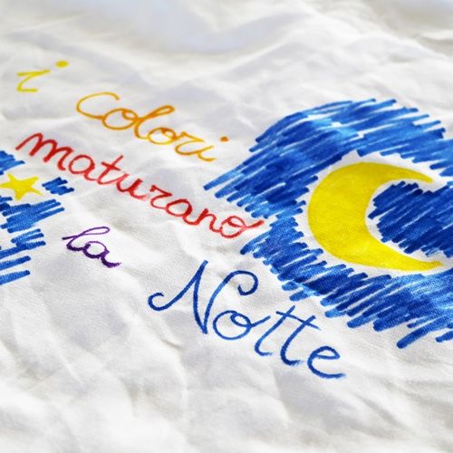 Close-up van wit textiel met een tekening van een nachtelijke hemel met maan en sterren, en de tekst 'I colori maturano la Notte', gemaakt met textielstiften.