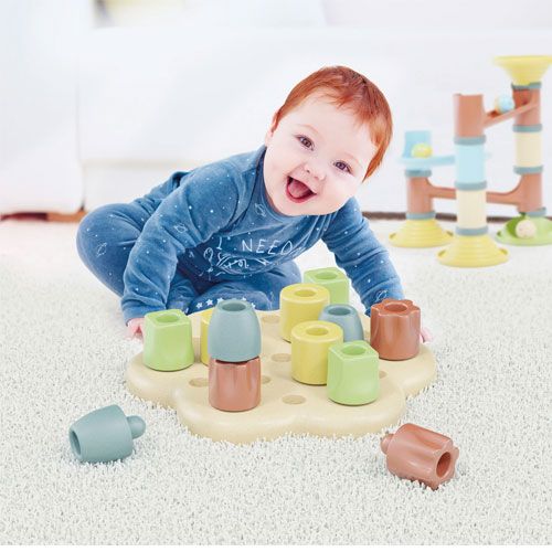 Een lachende baby speelt op een wit tapijt met het Quercetti Playbio sorteerspel met kleurrijke chunky peggs.
