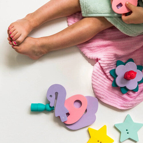 Kinderbenen naast een bad met de quut badpuzzel op de grond, inclusief schuimfiguren van een zwaan, bloem en sterren.