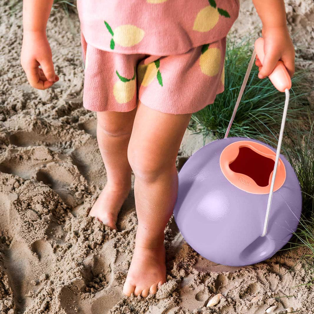 Een kind met roze kleding loopt op het zand en draagt de paarse balvormige strandemmer.
