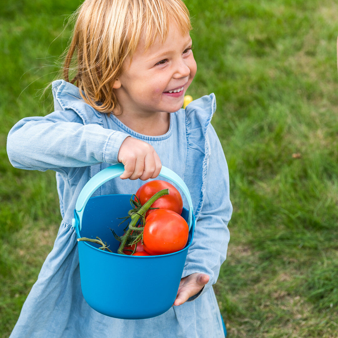 Lachend meisje in een spijkerjurk draagt een Quut blauwe emmer gevuld met tomaten.