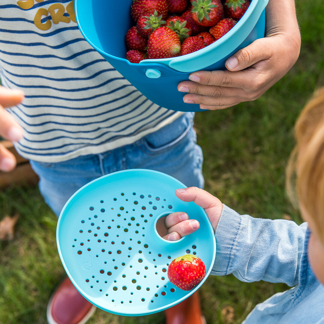 Kinderhanden houden een Quut blauwe emmer met aardbeien en een lichtblauwe zeef met een aardbei vast.
