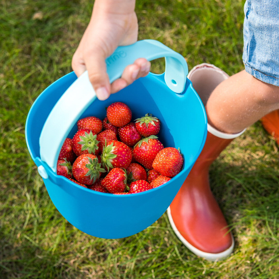 Een kind houdt een Quut blauwe emmer vol aardbeien vast, naast een rode laars op het gras.