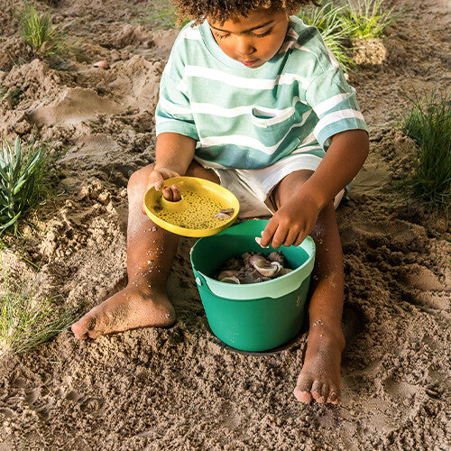 Een kind zit in het zand en sorteert schelpen met een gele zeef en groene emmer.