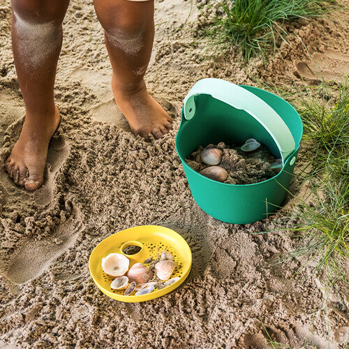 Blote kindervoeten in het zand naast een groene emmer en gele zeef met schelpen.