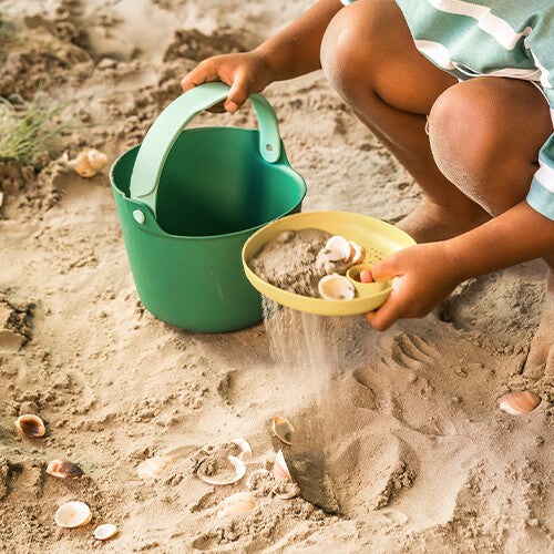 Een kind zeeft zand en schelpen met een gele zeef boven het zand.