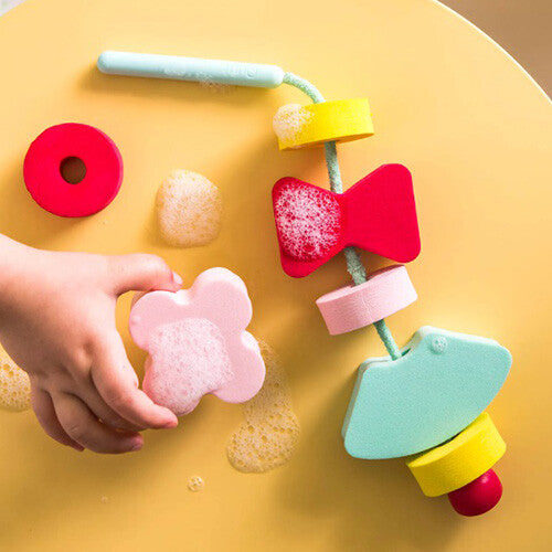 Een kinderhand houdt een roze vlindervormige schuimkraal van het quut badspeelgoed vast, bedekt met zeepsop.