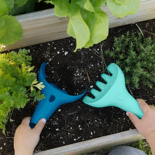Twee kinderhanden gebruiken de Quut blauwe schep en lichtblauwe hark in een moestuin.