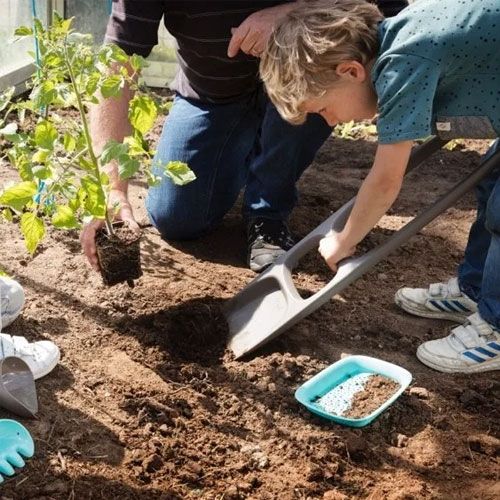 Kind en volwassene planten een plantje in de aarde, met een lichtblauwe Quut zeef voor zand of aarde op de grond.