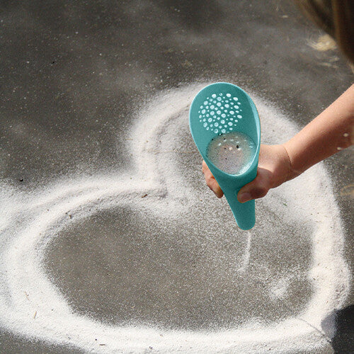 Een kinderhand strooit zand uit de lichtblauwe quut schep met zeef om een hart te vormen.