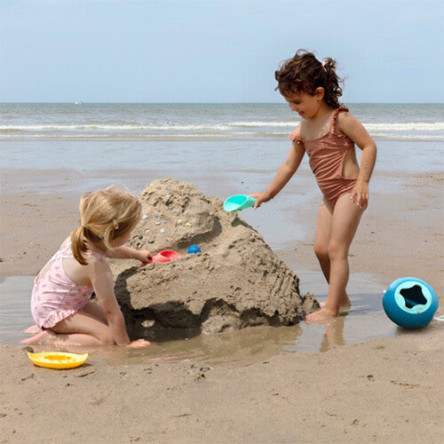 Twee kinderen spelen op het strand, één bouwt een zandkasteel met een quut schep.