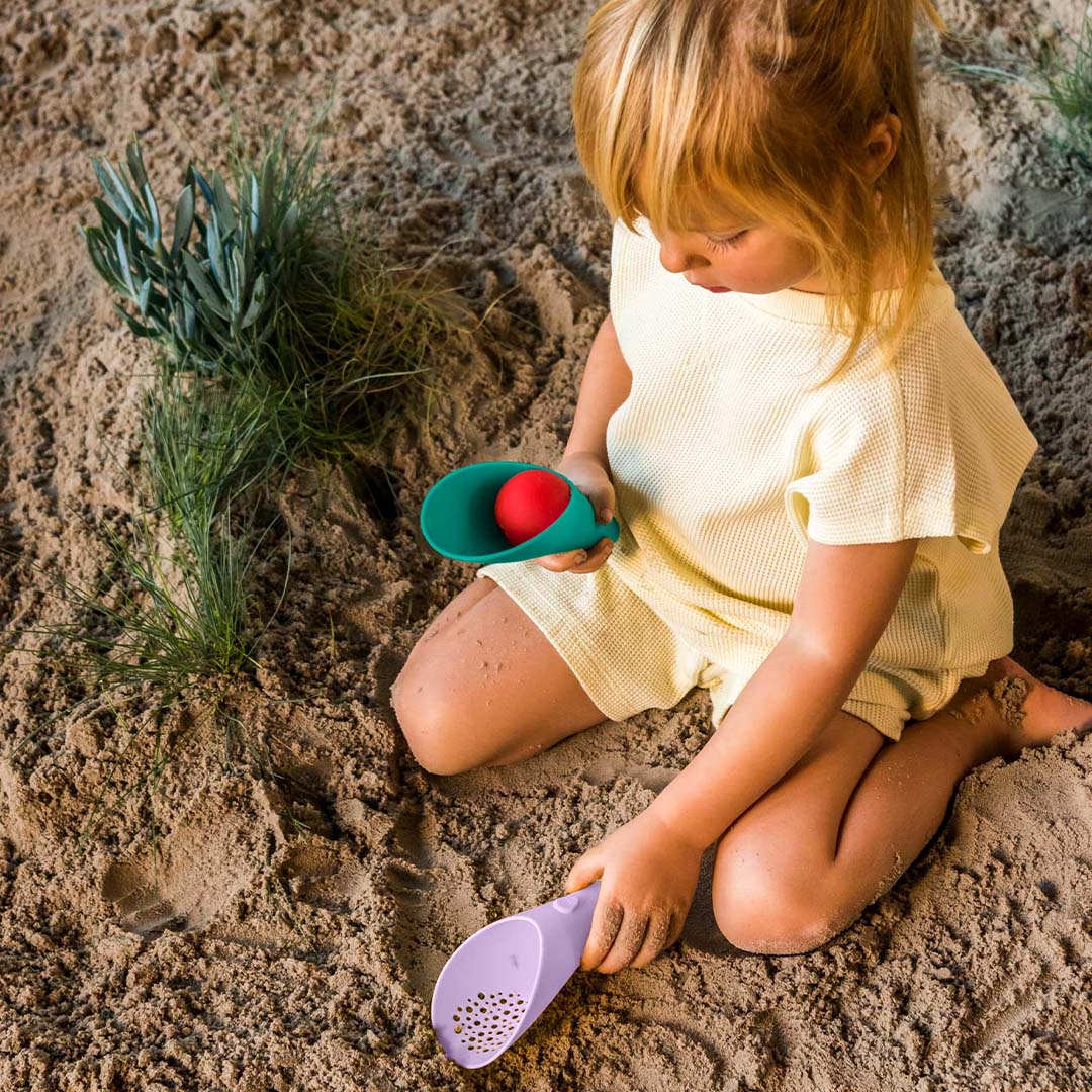 Een kind speelt in het zand met de Quut beach toys Cuppi set, bestaande uit een groene schep met rode bal en een paarse schep met zeef.